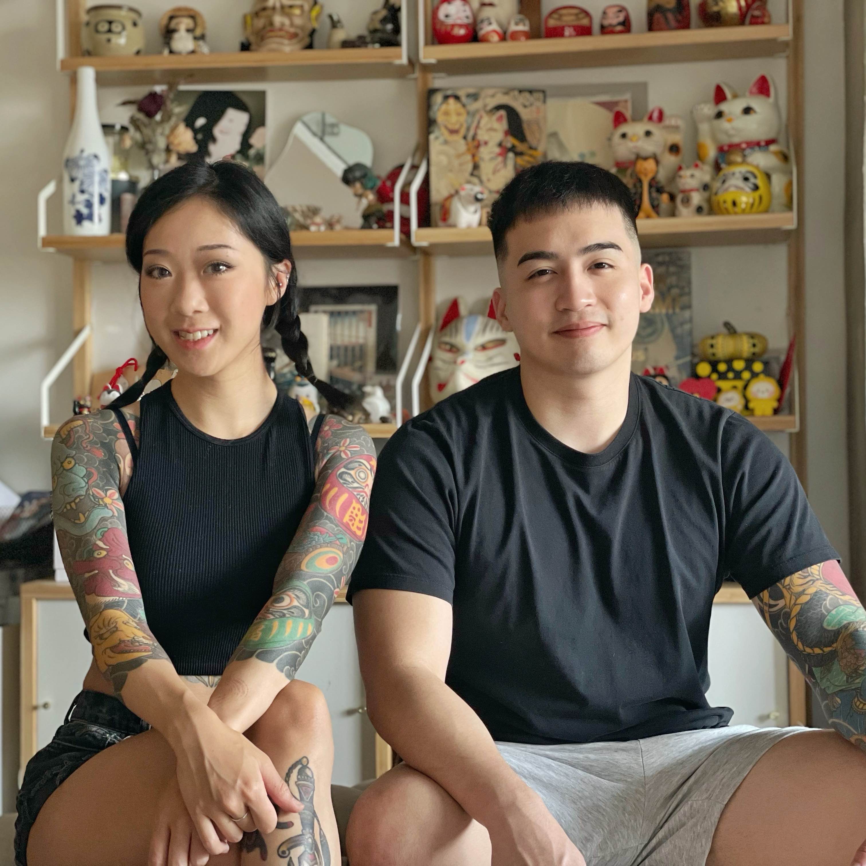Two tattoo artists sitting side by side in a studio space with shelves of lucky cat figurines behind them, both showing full sleeve tattoos and smiling at the camera.