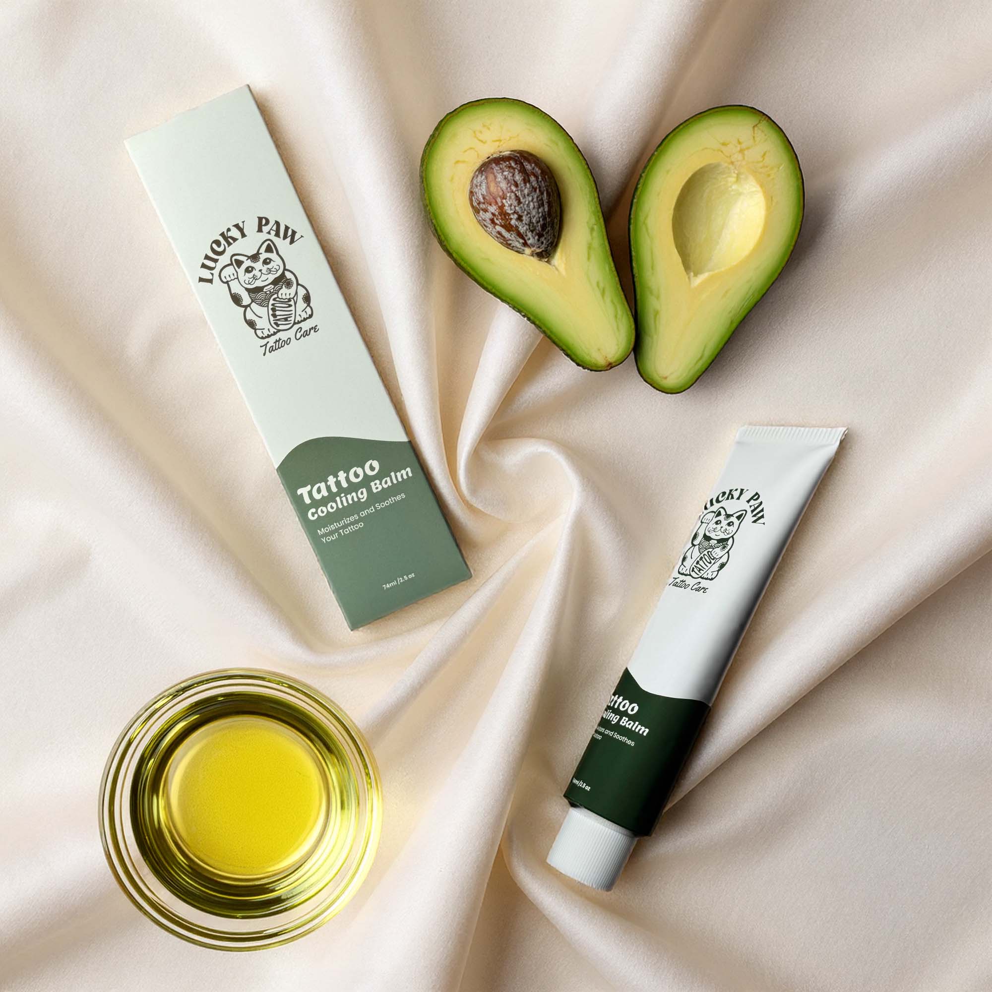 Lucky Paw Tattoo Cooling Balm tube and box arranged with halved avocado fruit and bowl of avocado oil on a beige satin fabric background.