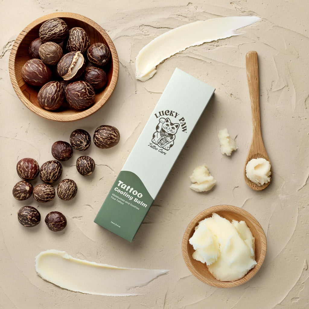 Flat lay of Lucky Paw Tattoo Cooling Balm packaging with bowls of shea butter, raw shea nuts, and balm texture on a beige surface.